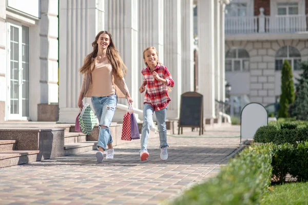 Усміхнена дівчина гуляє зі своєю мамою, несучи сумки на вулиці — стокове фото