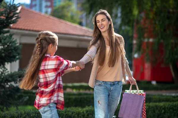 Девушка тянет свою улыбающуюся маму с пакетами для покупок на улице — стоковое фото