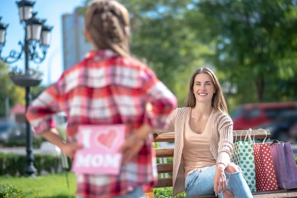 Πίσω όψη του κοριτσιού κρατώντας κάρτα για χαμογελαστή μαμά της κάθεται στο παγκάκι πάρκο — Φωτογραφία Αρχείου