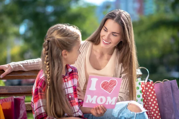 Hija sentada con su madre en el banco del parque, presentando su tarjeta —  Fotos de Stock