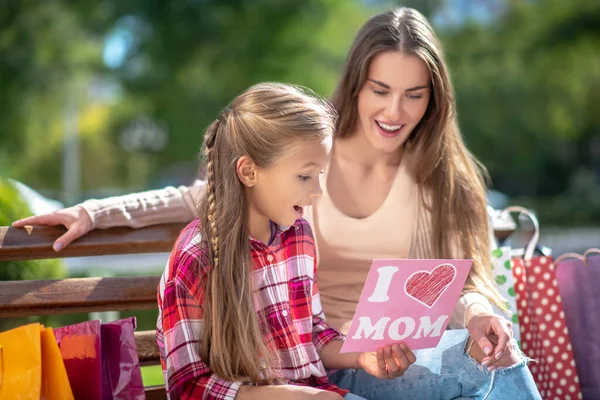 Hija sentada con su madre en el banco del parque, presentando su tarjeta —  Fotos de Stock