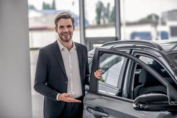 Sonriente moreno macho presentando coche nuevo, sosteniendo la puerta del coche —  Fotos de Stock