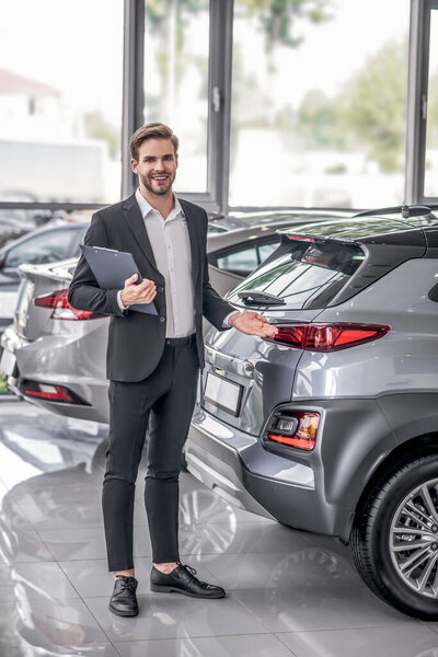 Bearded male holding clip folder, pointing at car boot in the showroom