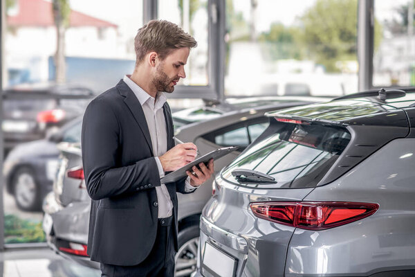 Bearded male examining car boot, taking notes in the showroom