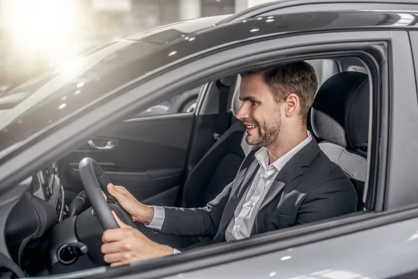 Sonriente joven macho sentado en el coche, sosteniendo el volante — Foto de Stock