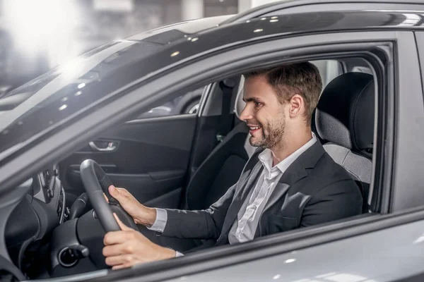 Sonriente joven macho sentado en el coche, sosteniendo el volante — Foto de Stock