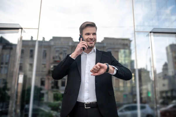 Elegantní mladý muž v černém obleku mluví do telefonu a dívá se na hodinky — Stock fotografie