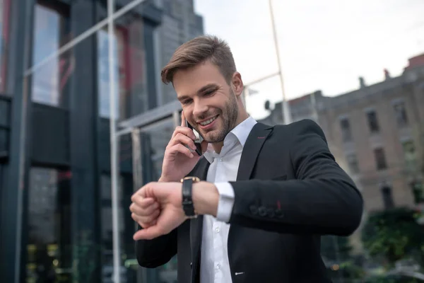 Jongeman in een zwart pak aan de telefoon en kijkend naar zijn horloge — Stockfoto
