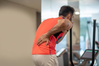 Parlak tişörtlü genç adam spor makinesinde egzersiz yapıyor ve sırt ağrısı çekiyor.