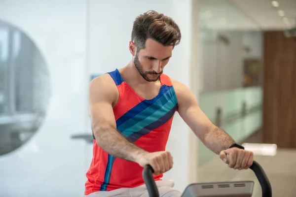 Молодой человек в яркой футболке, тренирующийся на тренажере и выглядящий сосредоточенным — стоковое фото