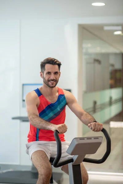 Hombre joven en camiseta brillante haciendo ejercicio en una máquina de gimnasio y sonriendo —  Fotos de Stock