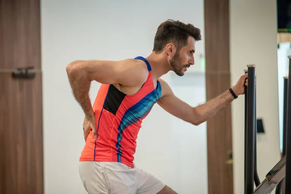 Man in helder tshirt oefenen op een fitnessapparaat en pijn in de rug voelen — Stockfoto