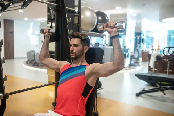 Jonge donkerharige man werkt aan zijn armen op een fitnessapparaat en ziet er geconcentreerd uit — Stockfoto