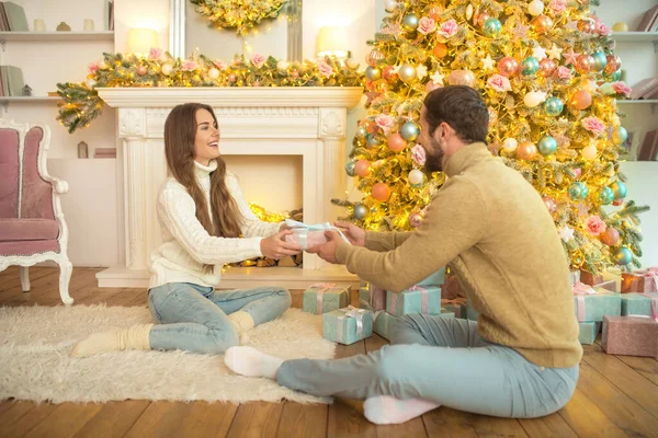 Mladý šťastný pár sedí v blízkosti vánoční strom a výměnu dárky — Stock fotografie