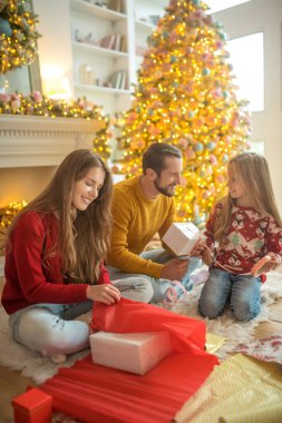 Genç bir aile yerde oturuyor ve Noel hediyelerini paketliyor.