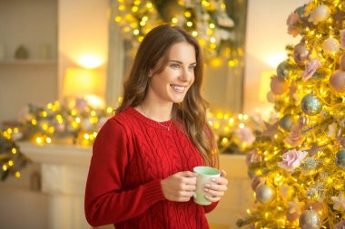 Koyu saçlı genç bir kadın elinde fincanla ayakta duruyor ve gülümsüyor.