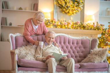Yaşlı bir adam elinde kitapla kanepede oturuyor ve karısı yanında duruyor.