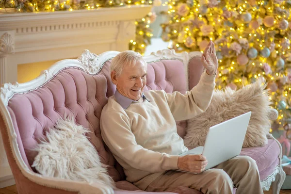 Grey-haired elderly man sitting on the sofa and having a video chat