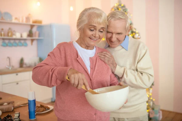 Äldre par står i köket, kvinna rör om något i en skål och ler — Stockfoto