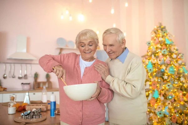 Couple âgé debout dans la cuisine, femme remuant quelque chose dans un bol et regardant satisfait — Photo
