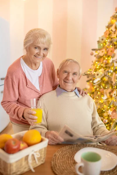 Anciano sentado en la mesa mientras su esposa le da jugo de naranja — Foto de Stock
