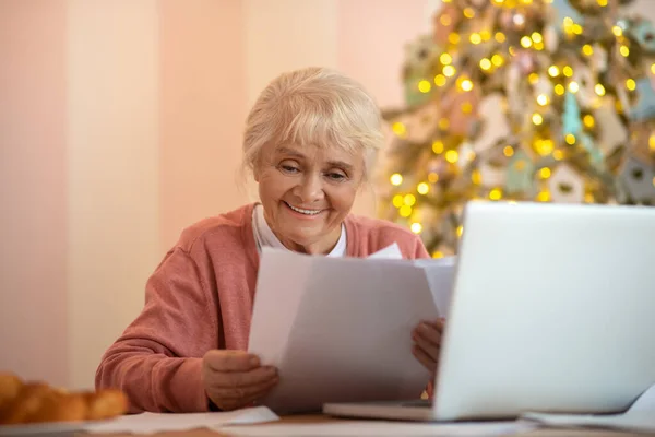 Пожилая женщина сидит за ноутбуком и читает газеты — стоковое фото