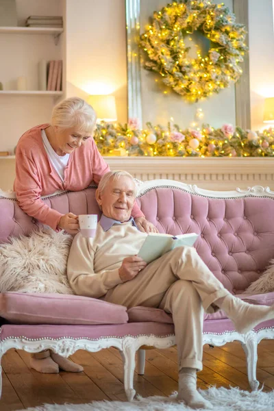 Idős férfi ül a kanapén egy könyvvel, és a felesége ad neki egy csésze teát. — Stock Fotó