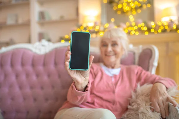 Starsza uśmiechnięta kobieta siedzi na kanapie i pokazuje swój smartfon — Zdjęcie stockowe