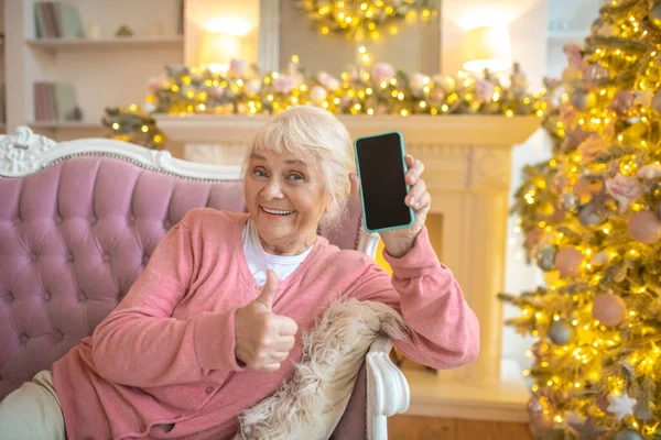 Starsza kobieta siedzi na kanapie i pokazuje swój smartfon — Zdjęcie stockowe