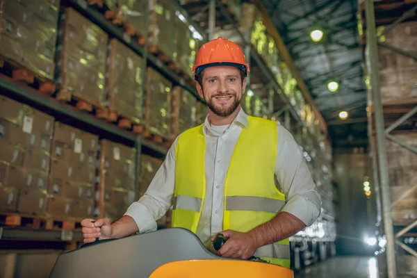 Trabajador de almacén en casco naranja de pie cerca de estantes con contenedores y sonriendo — Foto de Stock