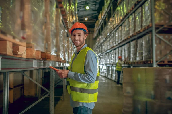 Hombre trabajador de almacén en casco naranja de pie cerca de contenedores y sosteniendo una tableta — Foto de Stock