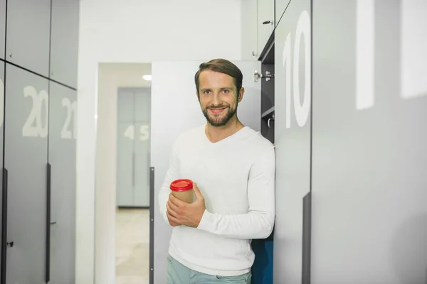 Young bearded man standing near the locker and holding cup with coffee — Stock Photo, Image