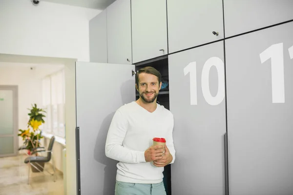 Young bearded man standing near the locker and holding cup with coffee — Stock Photo, Image