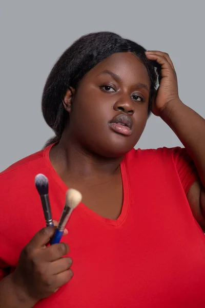Jovem mulher de pele escura em roupas vermelhas segurando escovas para maquiagem. — Fotografia de Stock