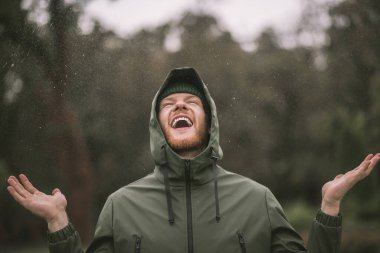 Young man in a green coat standing in the rain and smiling clipart