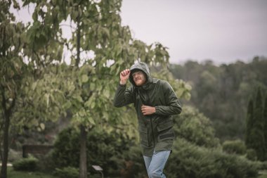 Yağmurun altında yürüyen kapüşonlu genç sakallı adam