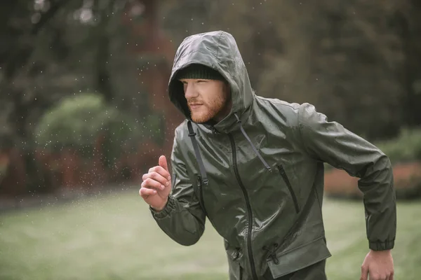 Młody brodaty mężczyzna w kapturze biegający pod deszczem — Zdjęcie stockowe