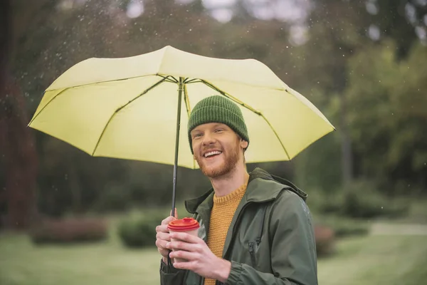 Young bearded man under yellow umbrella having coffee