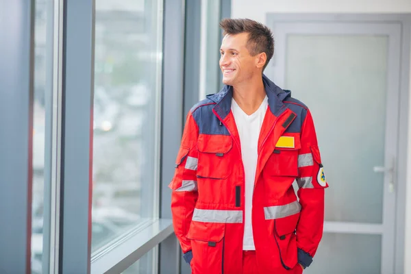 Médico varón de uniforme rojo de pie cerca de la ventana y sonriendo — Foto de Stock