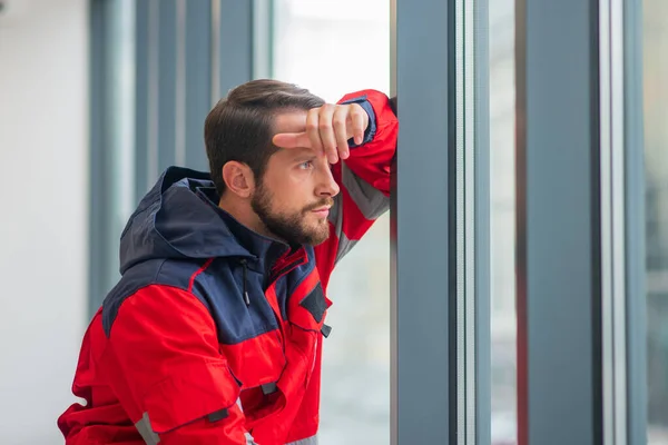 Médico masculino en uniforme rojo de pie cerca de la ventana y mirando reflexivo — Foto de Stock