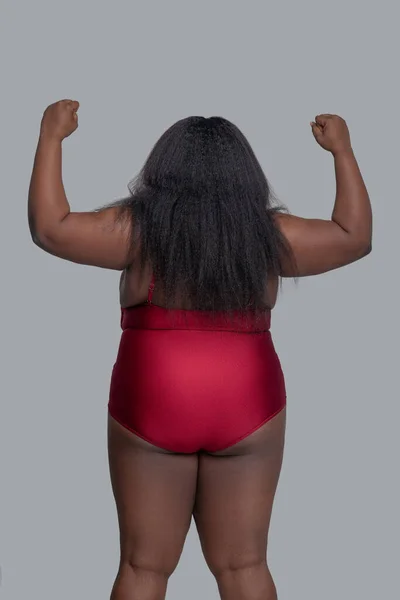 Afrikai amerikai fiatal nő piros alsóneműben, háttal áll, felemeli a kezét, és komolyan néz ki. — Stock Fotó