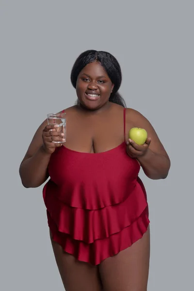 Dark-skinned smiling woman holding an apple and a glass of water — Stock Photo, Image