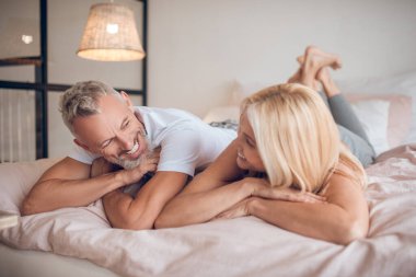 Gri saçlı bir adam ve sarışın bir kadın yatakta uzanıp birbirlerine bakıyorlar.