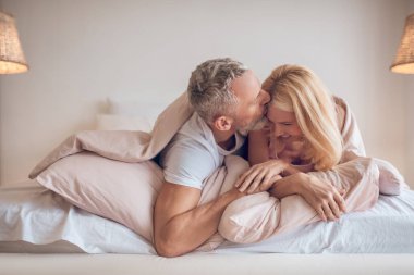 Gri saçlı bir adam ve sarışın bir kadın yatakta uzanıyor ve romantik görünüyorlar.