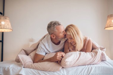 Gri saçlı bir adam ve sarışın bir kadın yatakta uzanıyor ve romantik görünüyorlar.