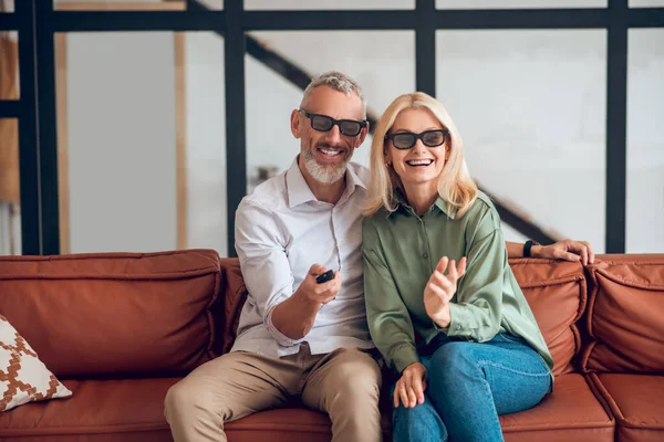 Зрелая пара в солнечных очках, сидящая на диване и включающая телевизор — стоковое фото