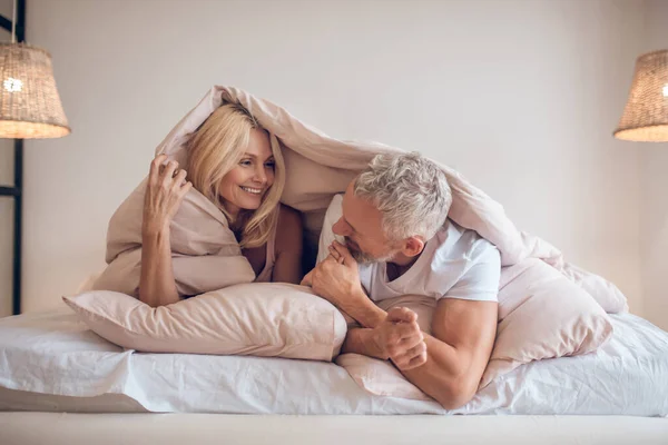Grey-haired man and a blonde woman lying in bed and having fun — Stock Photo, Image