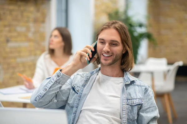 Jonge man met blond haar zit achter de laptop en praat aan de telefoon. — Stockfoto