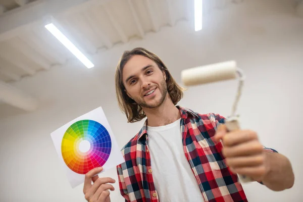 Joven hombre sonriente sosteniendo un rodillo de pintura y una paleta de colores — Foto de Stock