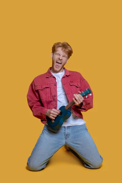 Man in red jacket playing the ukulele and looking enjoyed — Stock Photo, Image